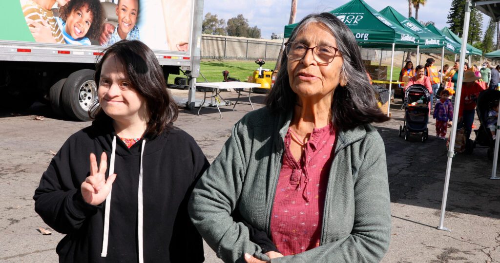 Two women stand outside at a mobile food pantry event. One, with long dark hair and a black hoodie, smiles and shows a peace sign. The other, with gray hair and glasses, wears a green sweater and red blouse. Tents and people are in the background.