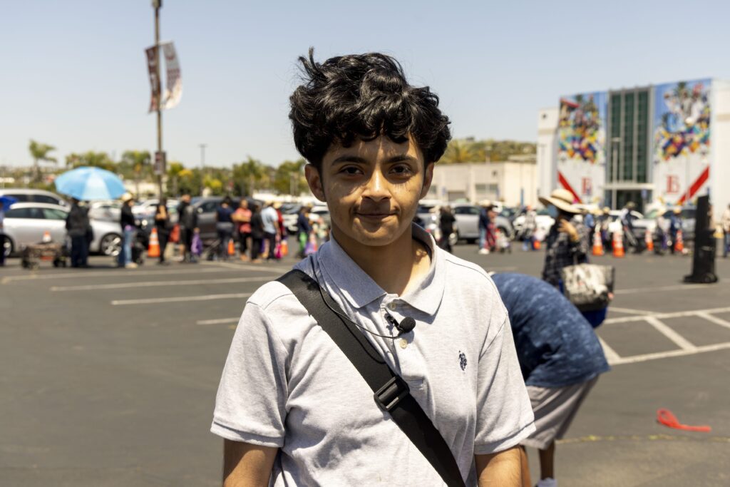 A young person with dark curly hair, wearing a light grey polo shirt and a black shoulder strap, stands in a sunny parking lot with a long line of people waiting at a mobile food pantry near a building in the background.