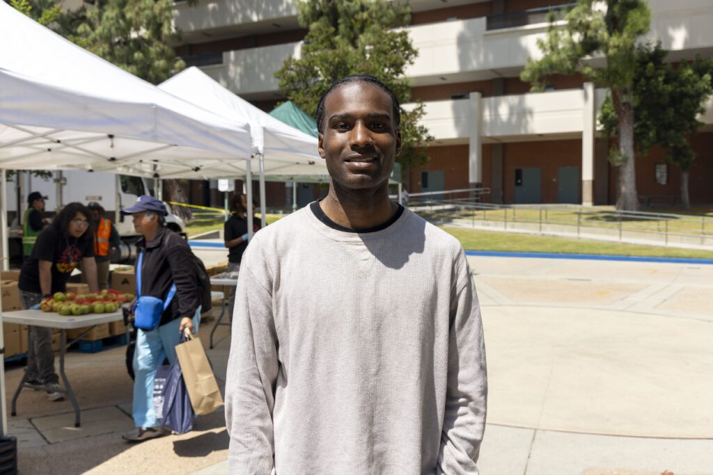 A man in a light gray long-sleeve shirt stands outdoors, smiling at the camera. Behind him, a mobile food pantry serves people under white tents, with tables of produce and a multi-story building framed by trees.