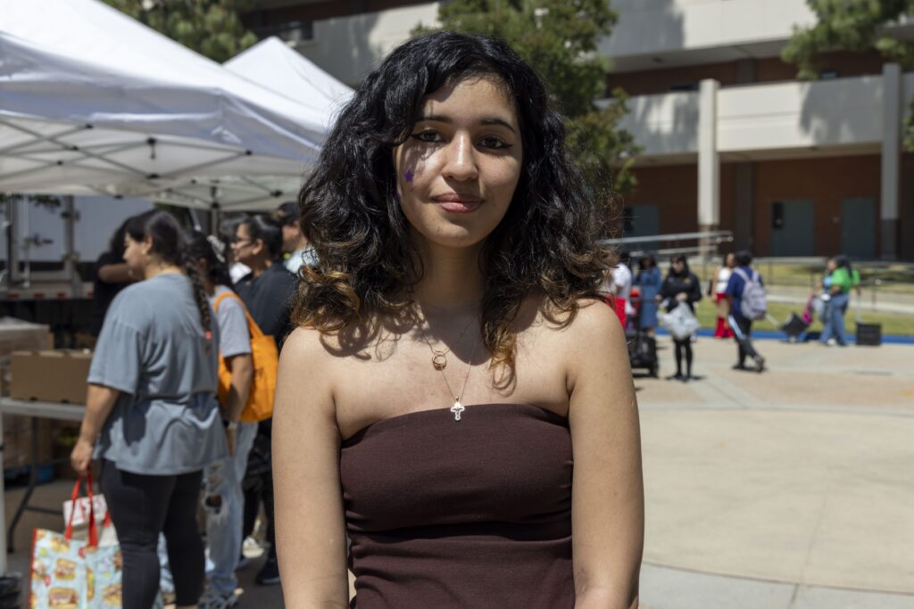 A young woman with wavy dark hair and a strapless brown top stands outdoors, smiling slightly. She has a purple marking on her left cheek. People, tents, and a mobile food pantry are visible in the background under sunny skies.
