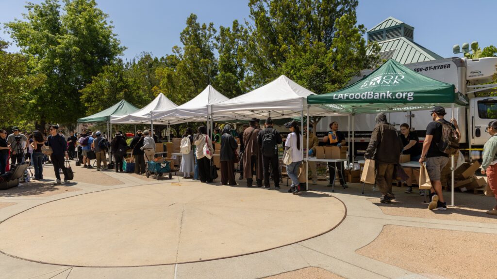 People line up under white and green tents at a mobile food pantry distribution event on a sunny day, with volunteers handing out food and supplies. Trees and a building are visible in the background.