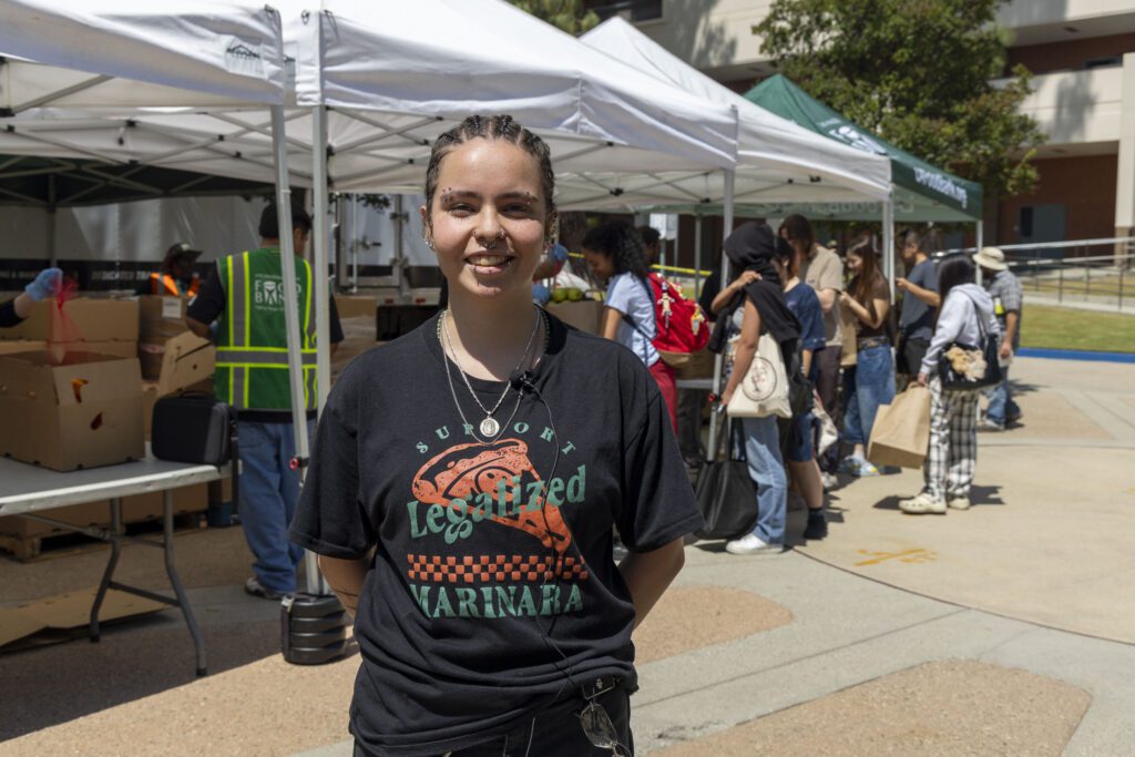 A person with braided hair smiles at the camera while standing outside near tents at a mobile food pantry. Several people are gathered in the background under the tents, some wearing backpacks and hats. It appears to be a sunny day.