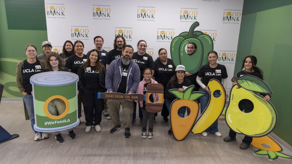 A group of people in UCLA Law shirts pose at a food bank under a banner that reads 