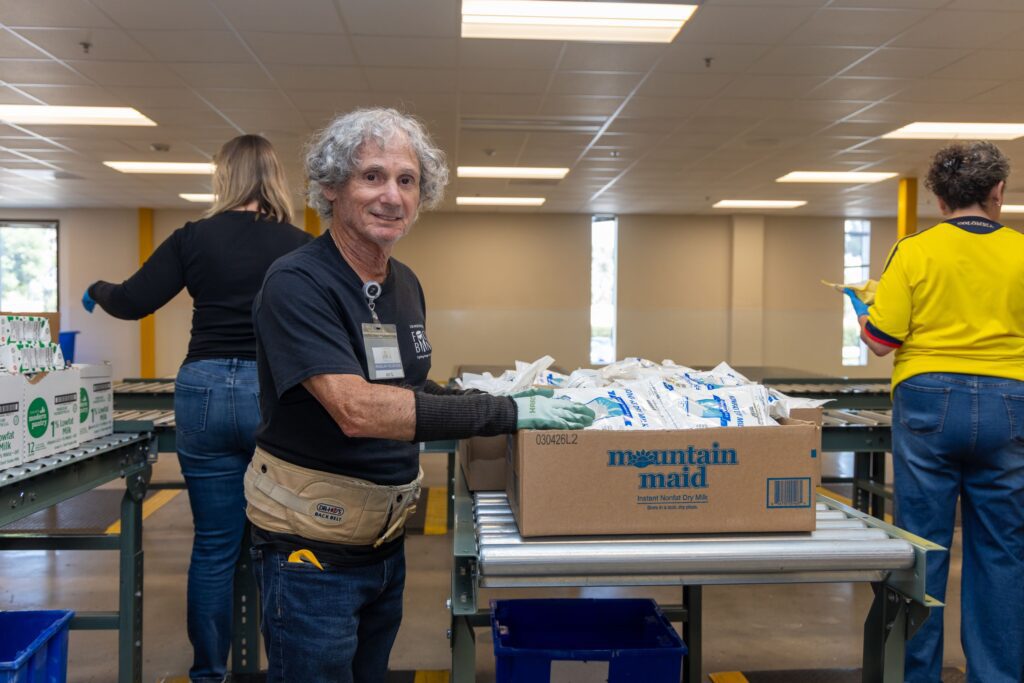 An older man with gray hair smiles while sorting packaged goods into a box labeled 