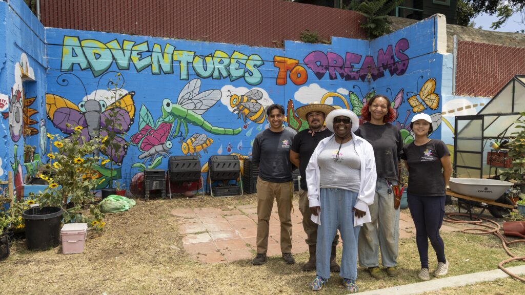 Five people stand smiling in front of a colorful mural that reads “Adventures to Dreams,” featuring cartoon insects and flowers, next to gardening equipment, sunflowers, and tables offering free food in an outdoor space.