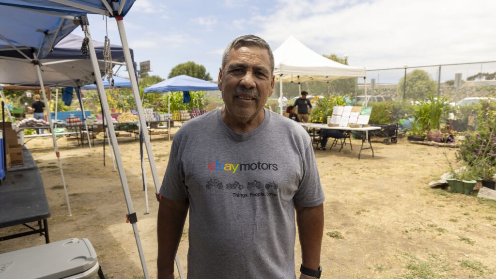 A man stands outdoors under blue and white tents, wearing a gray “eBay Motors” T-shirt at a mobile food pantry. Tables, plants, and people are visible in the background on a sunny day.