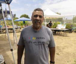 A man stands outdoors under blue and white tents, wearing a gray “eBay Motors” T-shirt at a mobile food pantry. Tables, plants, and people are visible in the background on a sunny day.