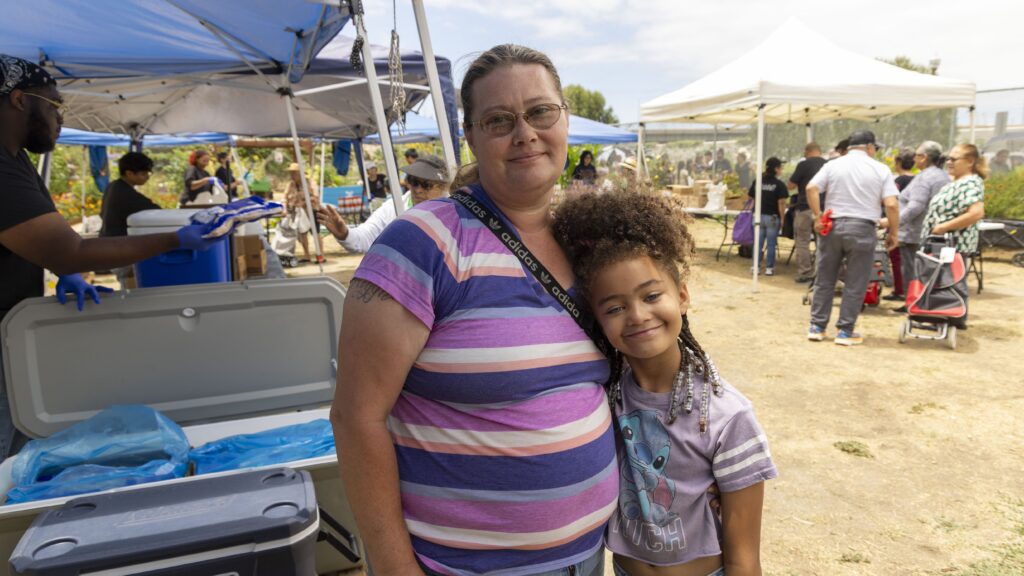 A woman and a young girl smile and hug at an outdoor event with free food, tents, people, and tables in the background on a sunny day.