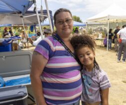 A woman and a young girl smile and hug at an outdoor event with free food, tents, people, and tables in the background on a sunny day.