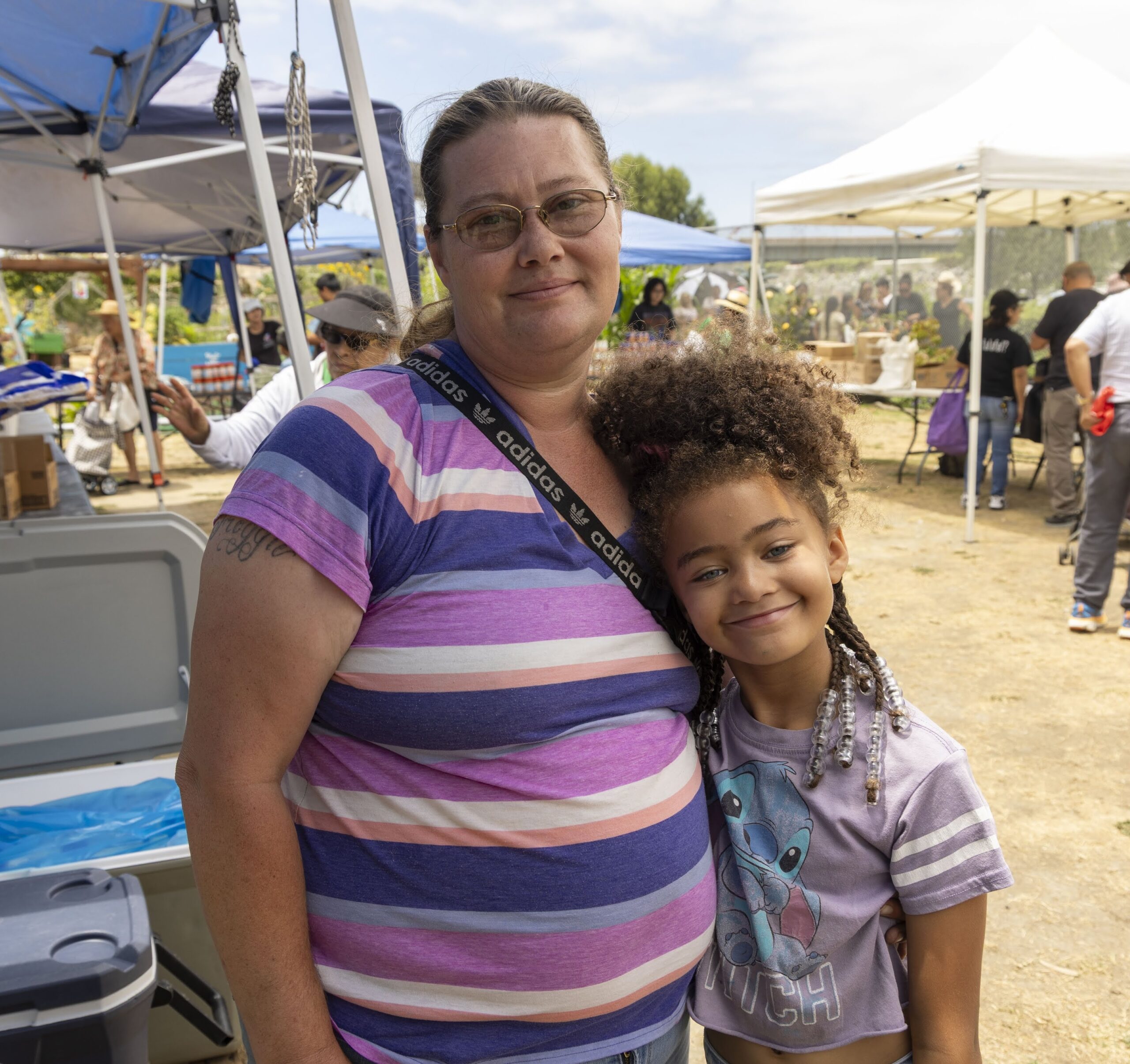 A woman in glasses and a striped shirt stands outdoors with a smiling young girl in a purple shirt. They are hugging at an event with tents, people, and free food in the background.