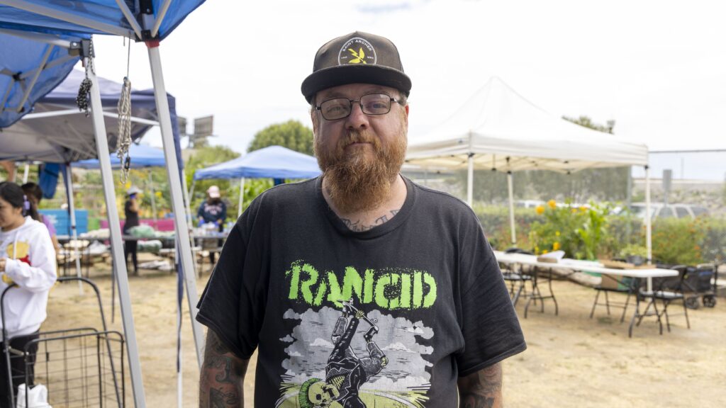 A bearded man wearing glasses, a black hat, and a “Rancid” T-shirt stands outdoors at a market with tents and tables in the background, possibly participating in food distribution activities.