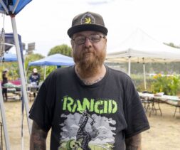 A bearded man wearing glasses, a black hat, and a “Rancid” T-shirt stands outdoors at a market with tents and tables in the background, possibly participating in food distribution activities.