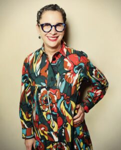 A person with glasses smiles while wearing a colorful, floral-patterned dress. In support of the Los Angeles Regional Food Bank, the backdrop is a soft, neutral tone as they stand confidently with one hand on their hip.
