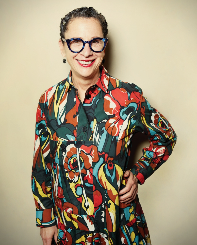 Nancy_Silverton.JPG A person with glasses smiles while wearing a colorful, floral-patterned dress. In support of the Los Angeles Regional Food Bank, the backdrop is a soft, neutral tone as they stand confidently with one hand on their hip.