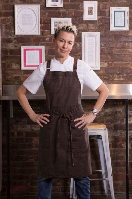 Elizabeth_Falkner_.JPEG A person stands confidently with hands on hips, wearing a white shirt and a brown apron, symbolizing their dedication to the Los Angeles Regional Food Bank. They are in front of a brick wall decorated with framed pictures, while a tall stool adds charm to the background.