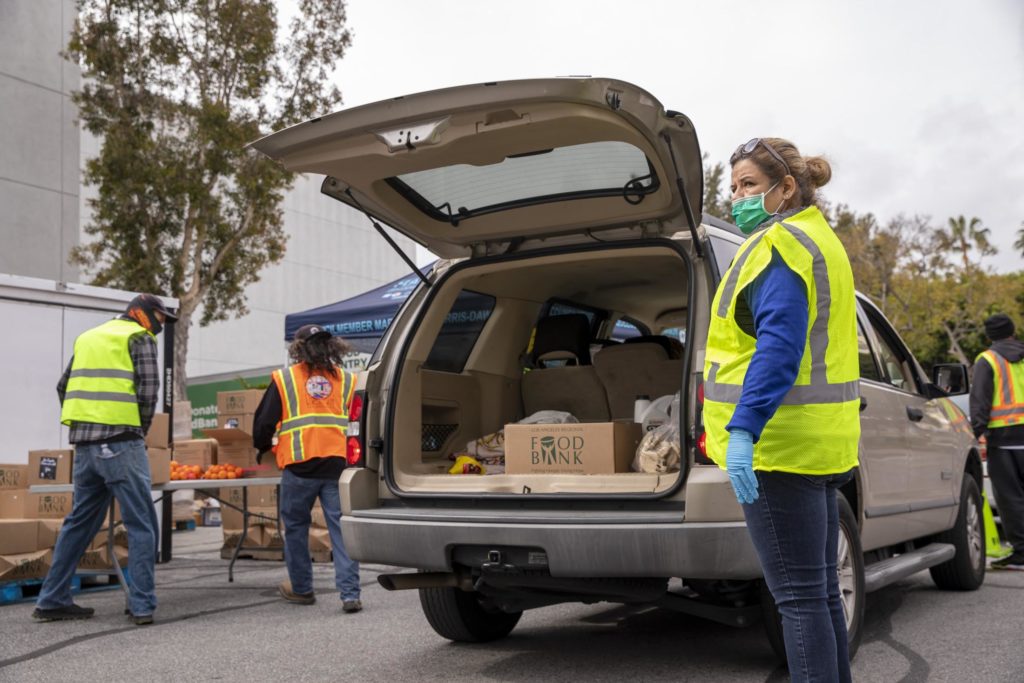 Volunteers in reflective vests and face masks distribute food at an outdoor Los Angeles Regional Food Bank event. An SUV with an open trunk is filled with boxes labeled "Food Bank." Nearby, tables are neatly organized with boxes and fresh produce.
