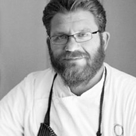 foodbank-michael-cimarusti-275x275-v1 A bearded man wearing glasses and a chef's jacket with an apron, smiling at the camera. The image is in black and white, capturing his dedication as he volunteers at the Los Angeles Regional Food Bank.