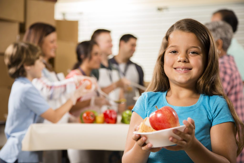 A smiling girl holds a bowl with an apple at a Los Angeles Regional Food Bank event. In the background, a group of people, including children and adults, are sorting and organizing food items on a table in this community setting.