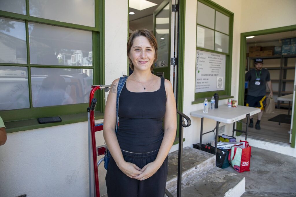 A woman in a black tank top stands smiling outside a building with green trim, possibly part of a local food bank. Behind her, a man works at a table with supplies, adding to the casual and friendly atmosphere.