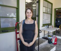 A woman in a black tank top stands smiling outside a building with green trim, possibly part of a local food bank. Behind her, a man works at a table with supplies, adding to the casual and friendly atmosphere.