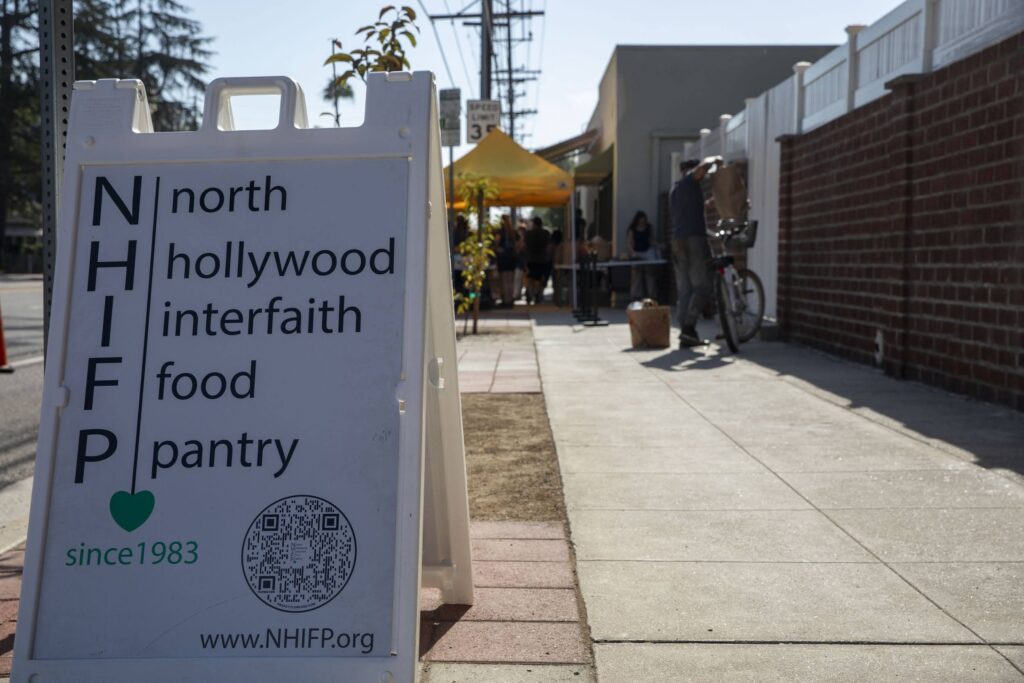 A sidewalk sandwich board reads “North Hollywood Interfaith Food Pantry” with a QR code and website; further down the sidewalk, people gather under a yellow tent at the food bank.