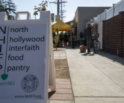 A sidewalk sandwich board reads “North Hollywood Interfaith Food Pantry” with a QR code and website; further down the sidewalk, people gather under a yellow tent at the food bank.