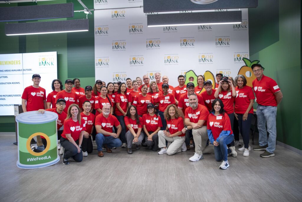 A group of people in matching red shirts pose and smile together at the Los Angeles Regional Food Bank, proudly representing the Los Angeles Dodgers Foundation. Some stand, others kneel, and there’s a large can prop with “#WeFeedLA” text visible in front.