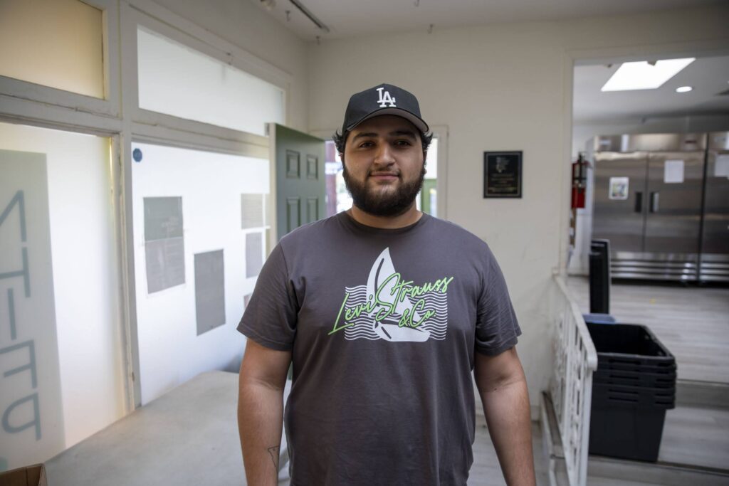 A man with a beard wearing a gray graphic t-shirt and a black LA cap stands indoors in a bright room, possibly at the North Hollywood Interfaith Food Pantry, with a green door and kitchen equipment visible in the background.