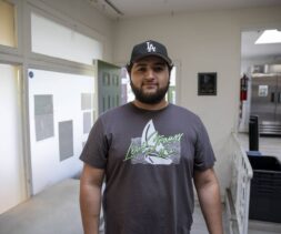 A man with a beard wearing a gray graphic t-shirt and a black LA cap stands indoors in a bright room, possibly at the North Hollywood Interfaith Food Pantry, with a green door and kitchen equipment visible in the background.