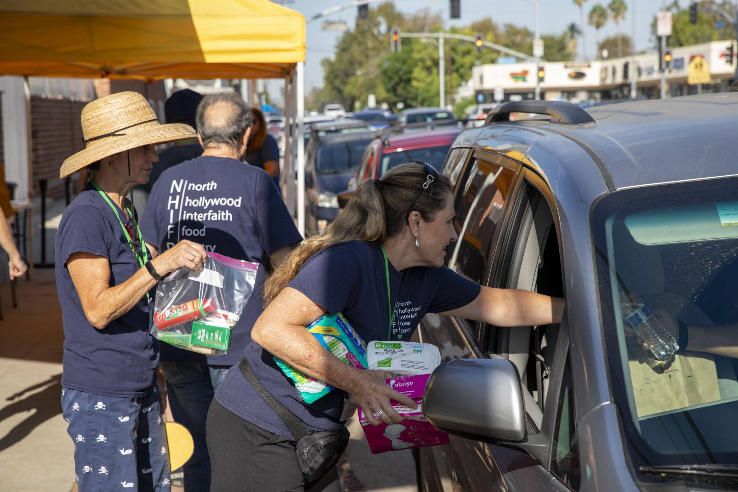 Volunteers in blue shirts from the North Hollywood Interfaith Food Pantry distribute food and supplies to people in cars at a drive-through donation event on a sunny day.