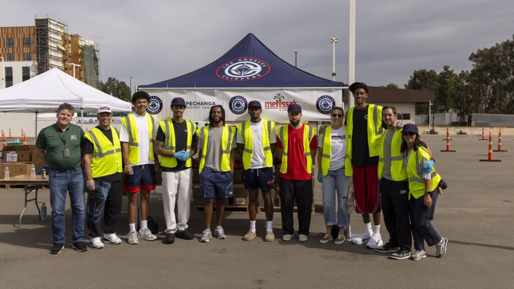 A group of ten people, some wearing Los Angeles Clippers hats and neon yellow Sodexo safety vests, pose for a photo in front of a branded canopy tent at an outdoor event.